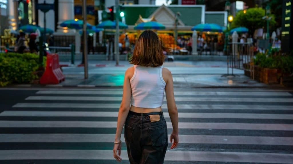 A woman in a white tank top walks across a city crosswalk away from the camera.