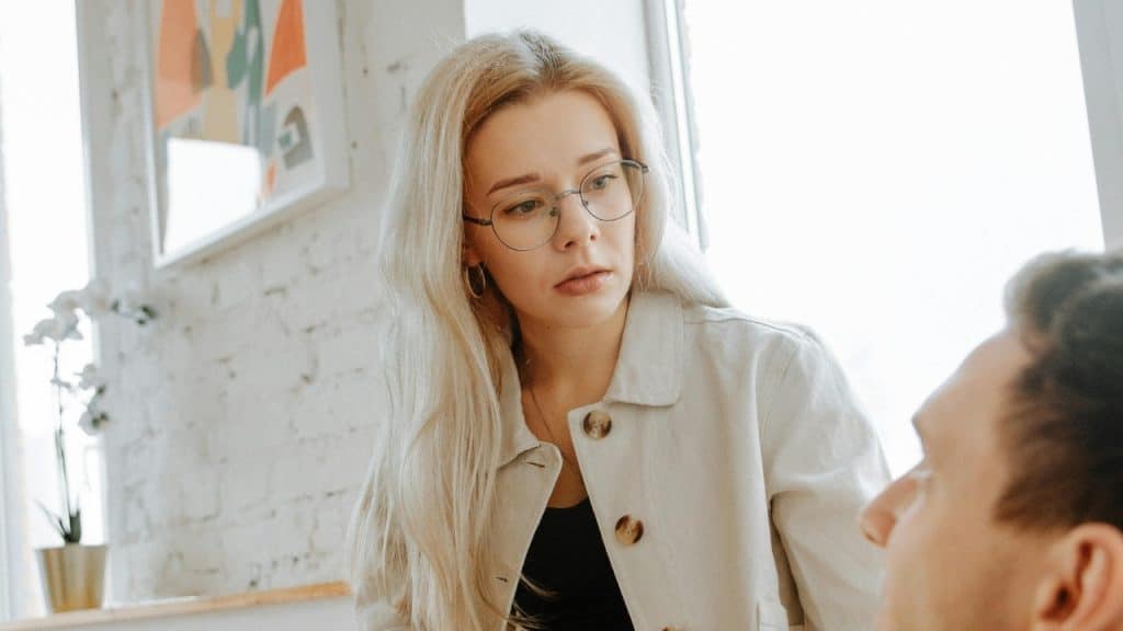 A blonde woman with glasses looks intently toward a manβs face in a bright room.