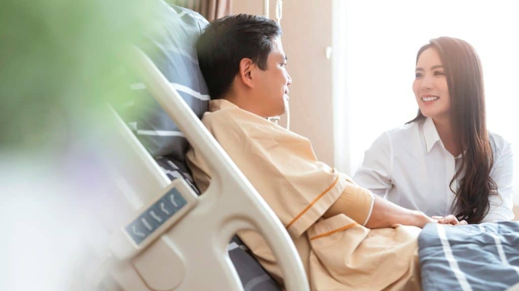 A woman in a white shirt smiles while sitting beside a man in a hospital bed.