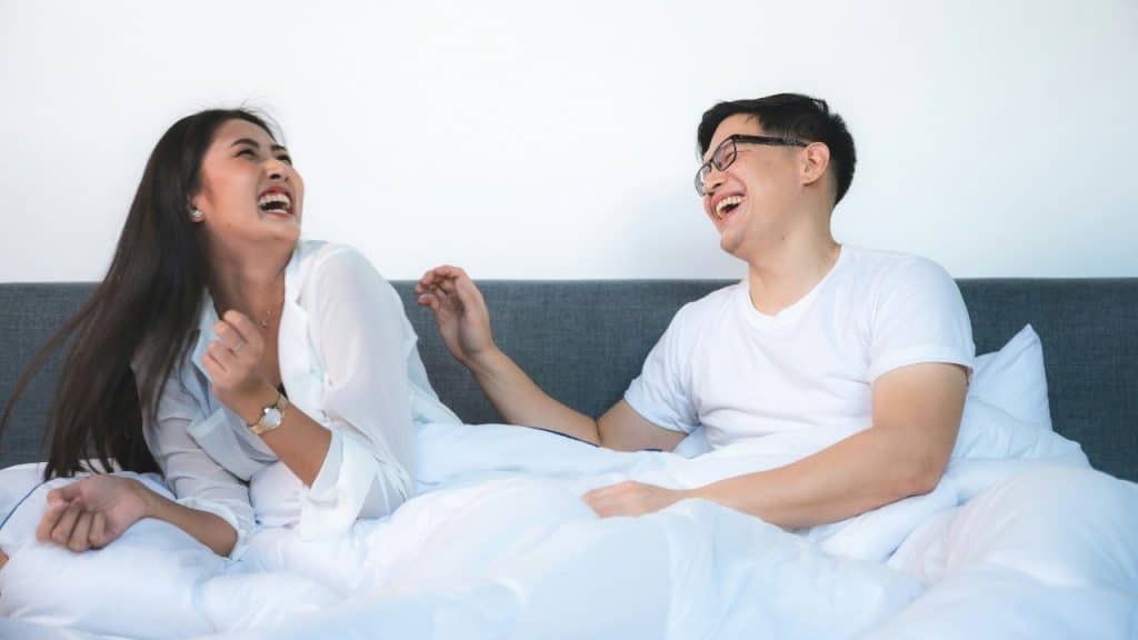 A man and woman laugh together while sitting in a bed with white pillows and sheets.
