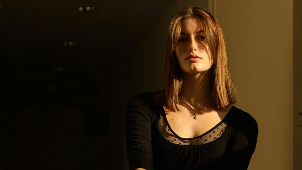 A woman with brown hair looks directly at the camera while standing in a dark hallway.