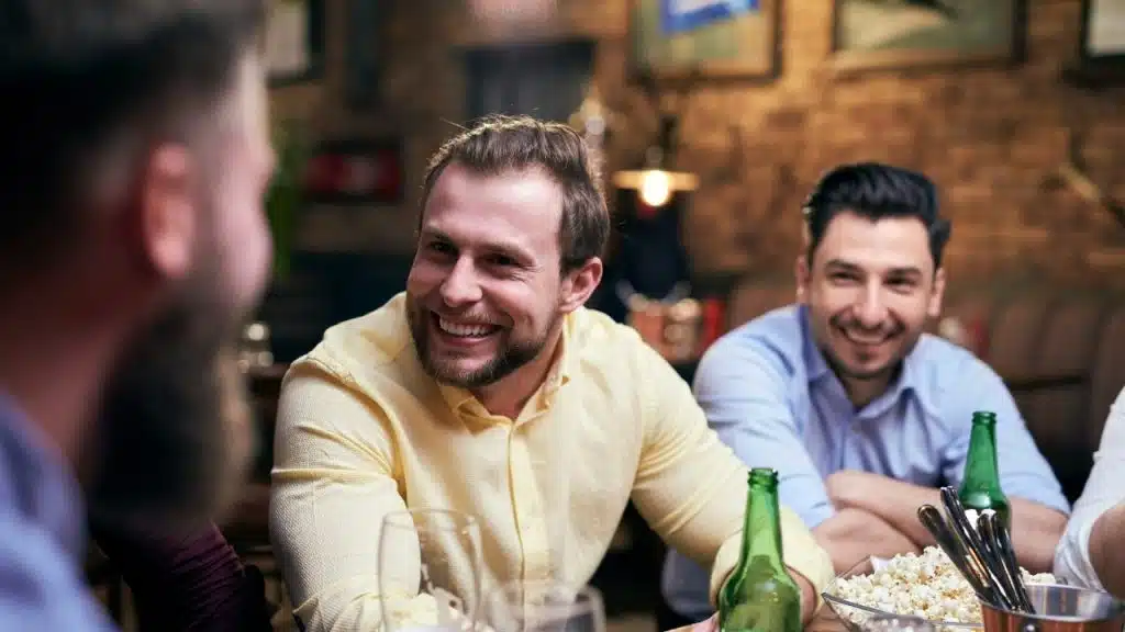 Three men sitting at a bar table with beer bottles and a bowl of popcorn.