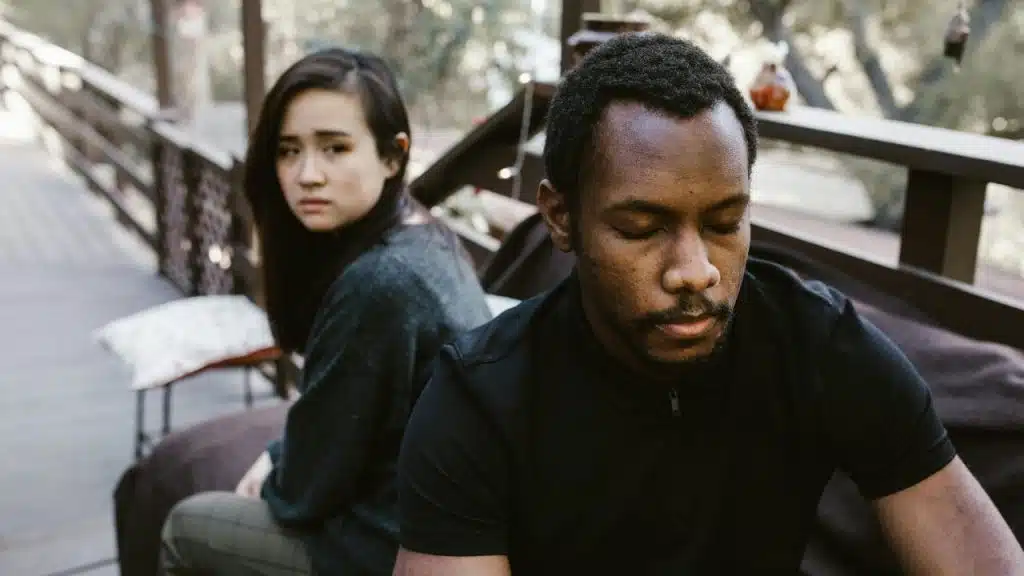 A man sits in the foreground with eyes closed while a woman looks on behind him.