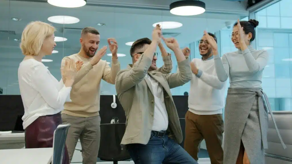 A group of people in an office setting are cheering and clapping with their arms raised.