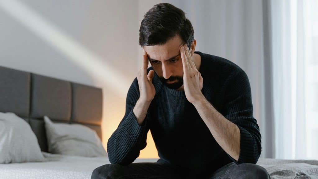 A man with a beard sits on a bed holding his head with both hands.