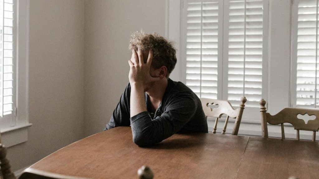 A person sits at a wooden table with their face buried in one hand.