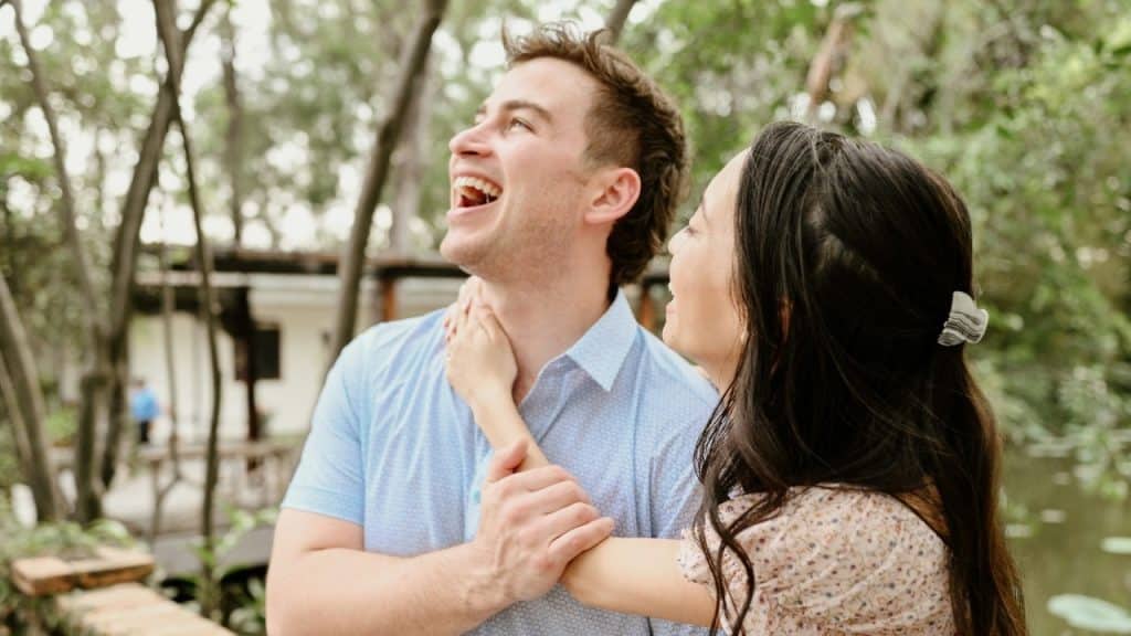 A man laughs loudly while a woman stands close to him in a wooded area.