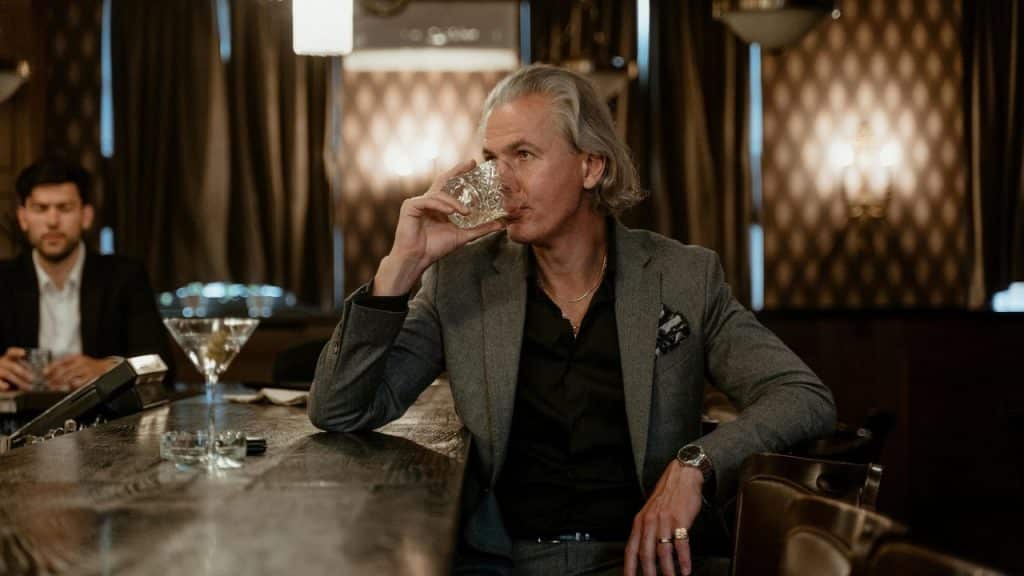 A man in a suit sits at a dark bar drinking from a crystal glass.