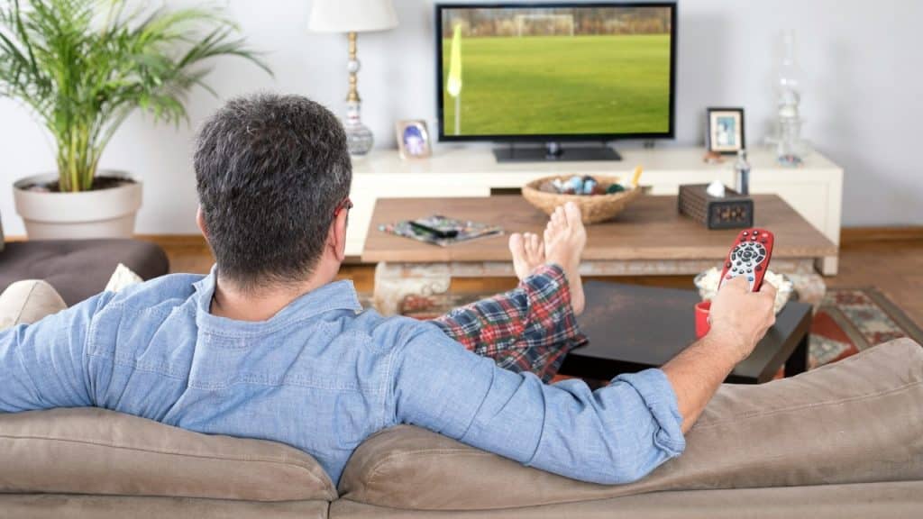 A man in pajamas watches a soccer game on television while holding a red remote.