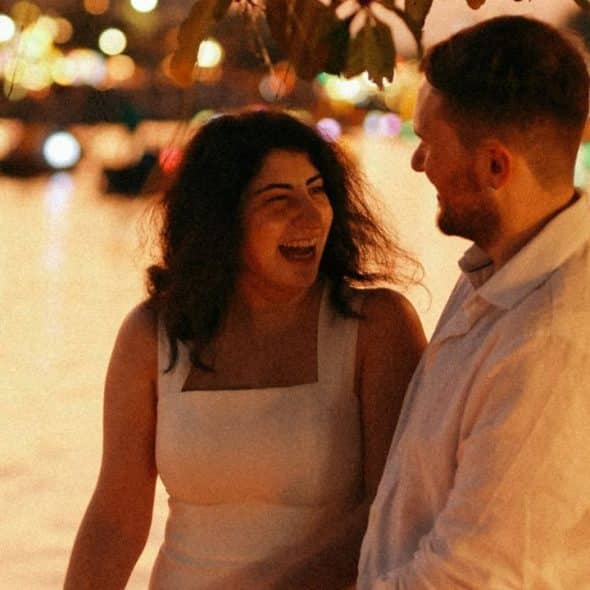 A man and woman laugh together outdoors at night beside a glowing body of water.