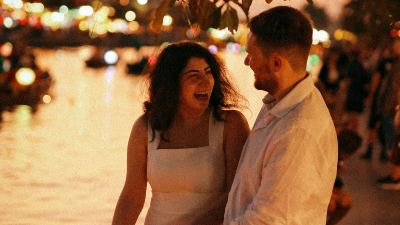 A man and woman laugh together outdoors at night beside a glowing body of water.