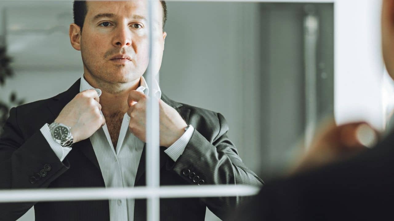 A man in a dark suit adjusts his white shirt collar while looking into a mirror.
