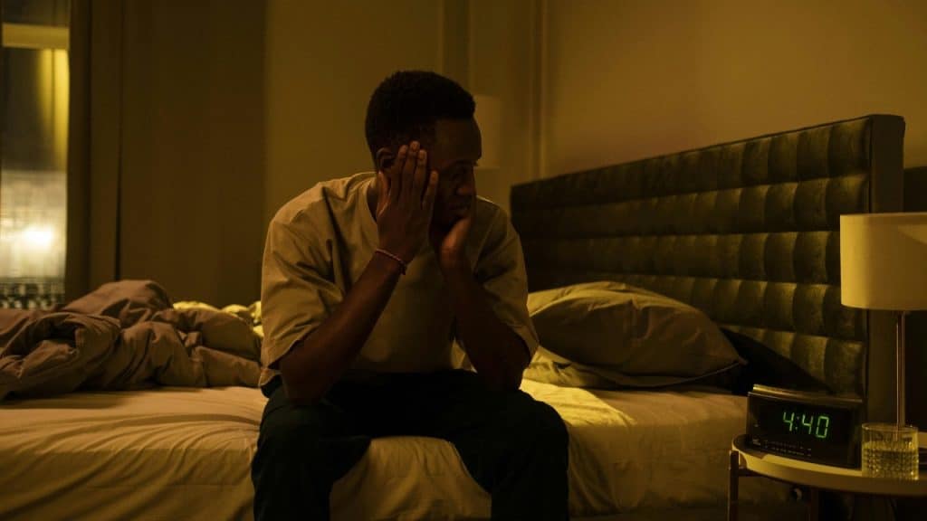 A man sits on the edge of a bed in a dark room holding his head.
