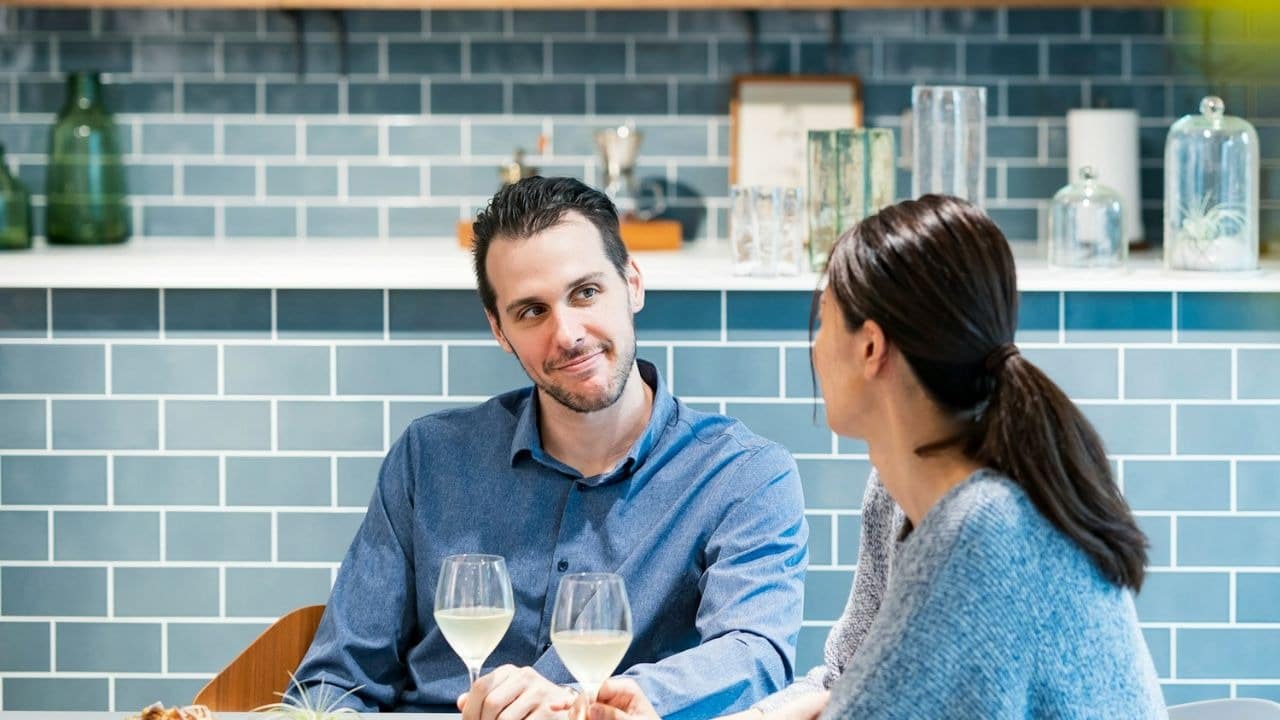 A man in a blue shirt looks at a woman while holding a glass of wine.