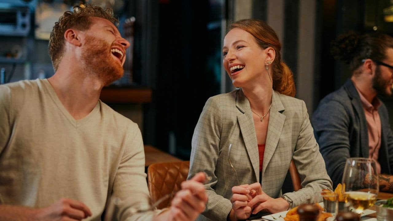 A man and woman laugh while sitting at a dinner table with food and drinks.