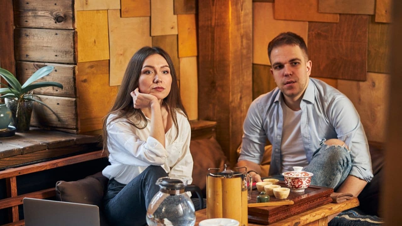 A man and woman sit near a wooden wall with tea sets on a table.