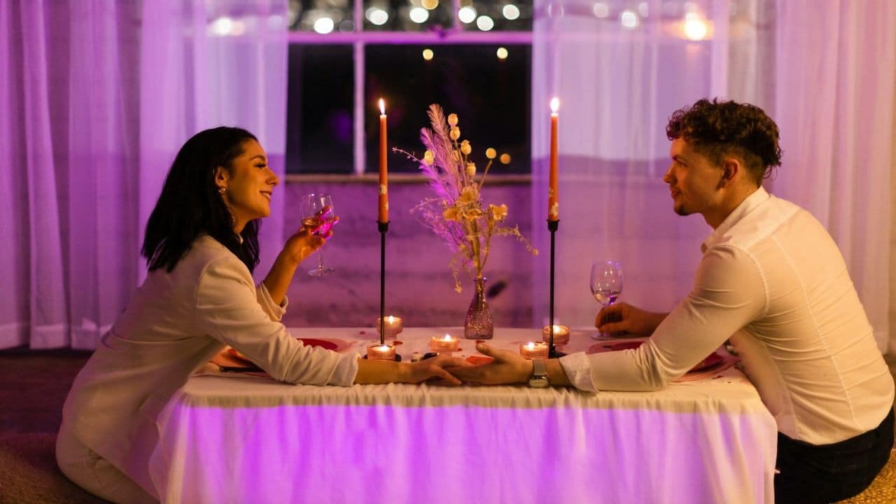 A man and woman hold hands at a candlelit table with wine under purple light.