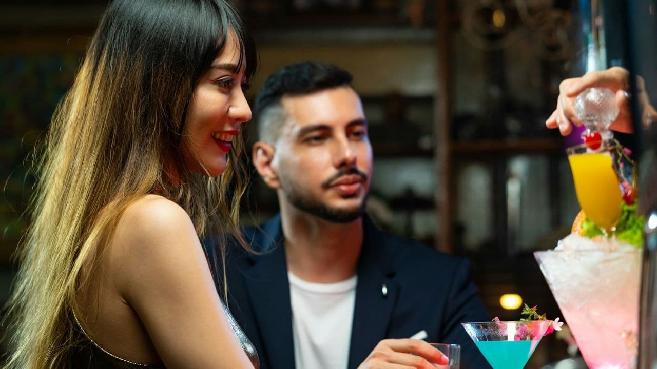 A woman and man watch a person prepare a colorful, layered drink at a bar.