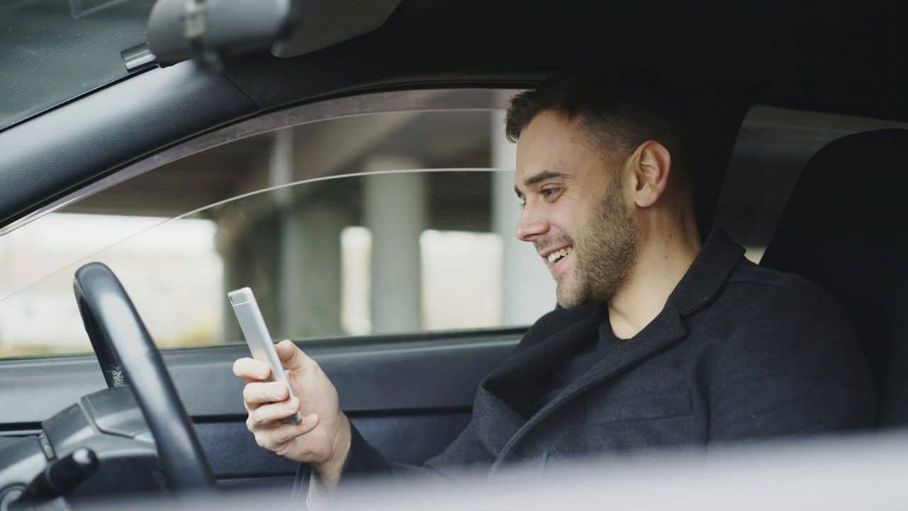 A smiling man sits in a car while looking at a mobile phone in his hand.