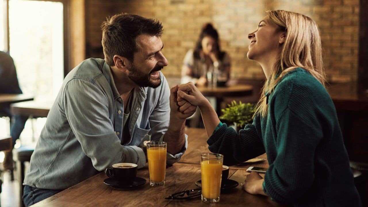 A man and woman hold hands and laugh while sitting at a cafe table.