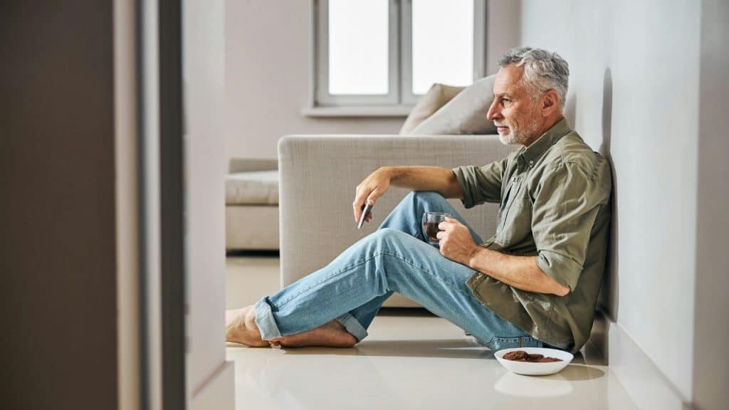 An older man sits on the floor leaning against a wall, holding a glass mug.