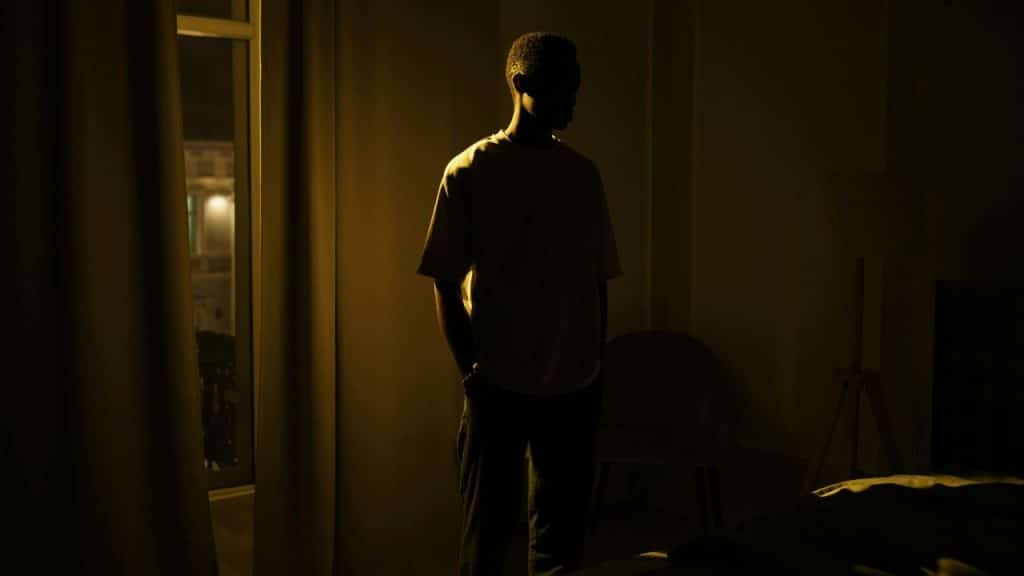 A man stands in a dark room with a single warm light source hitting his shoulder.