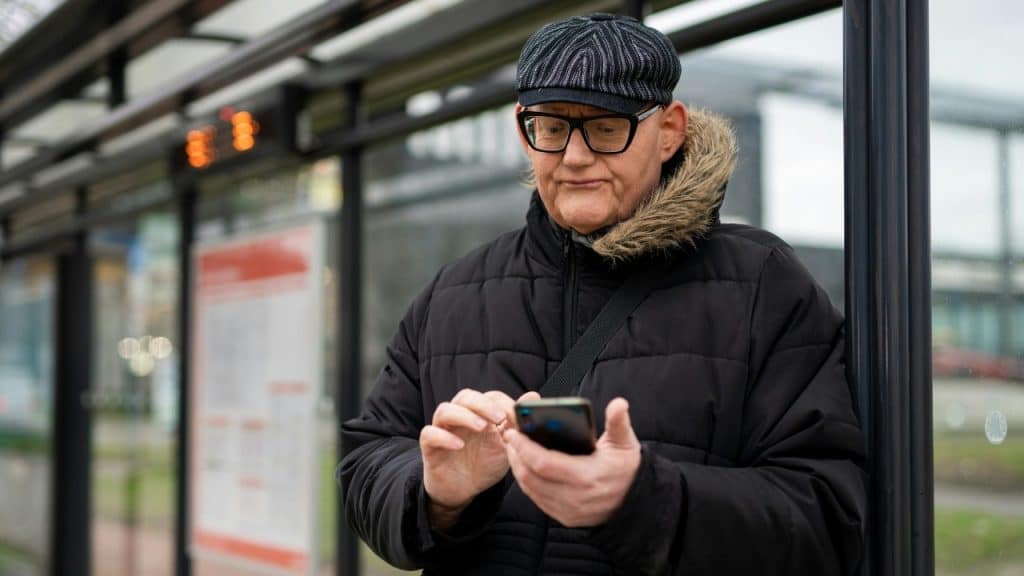 A man in a winter coat and flat cap looks down at a handheld mobile phone.
