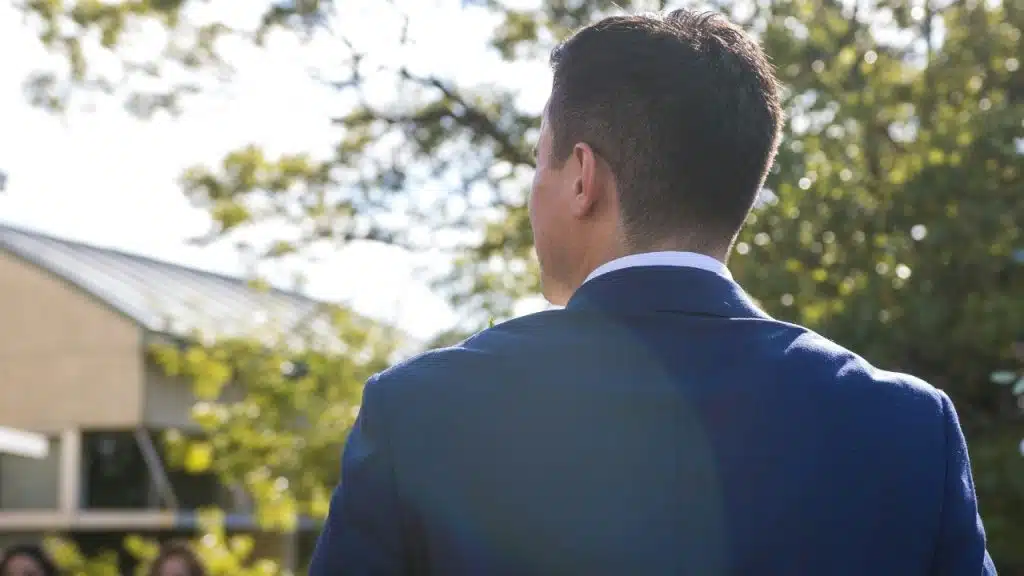 A man in a blue suit stands outdoors with his back to the camera.