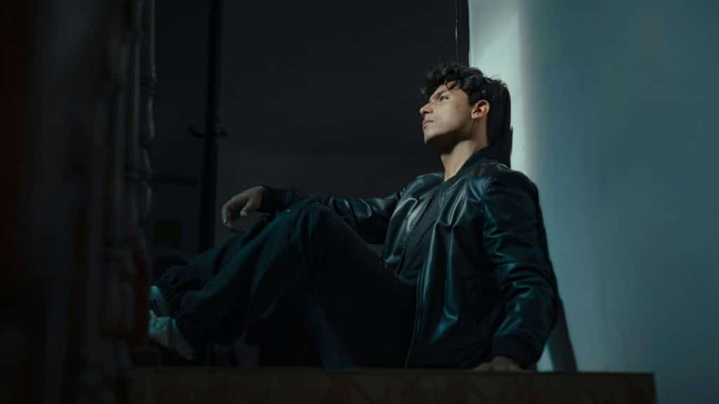 A man in a leather jacket sits on stairs, leaning against a white wall.