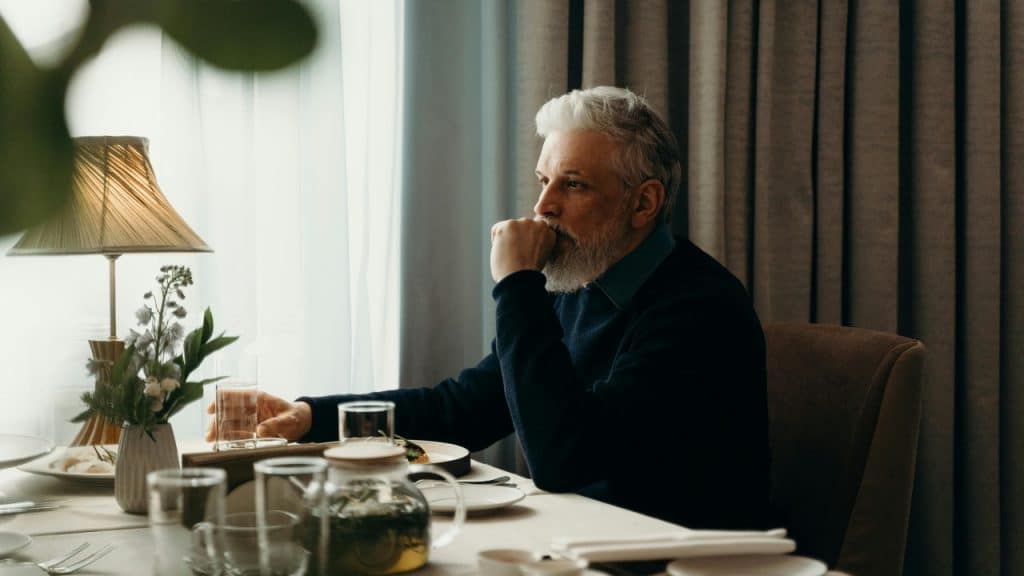 A bearded man with white hair sits alone at a set dining table indoors.