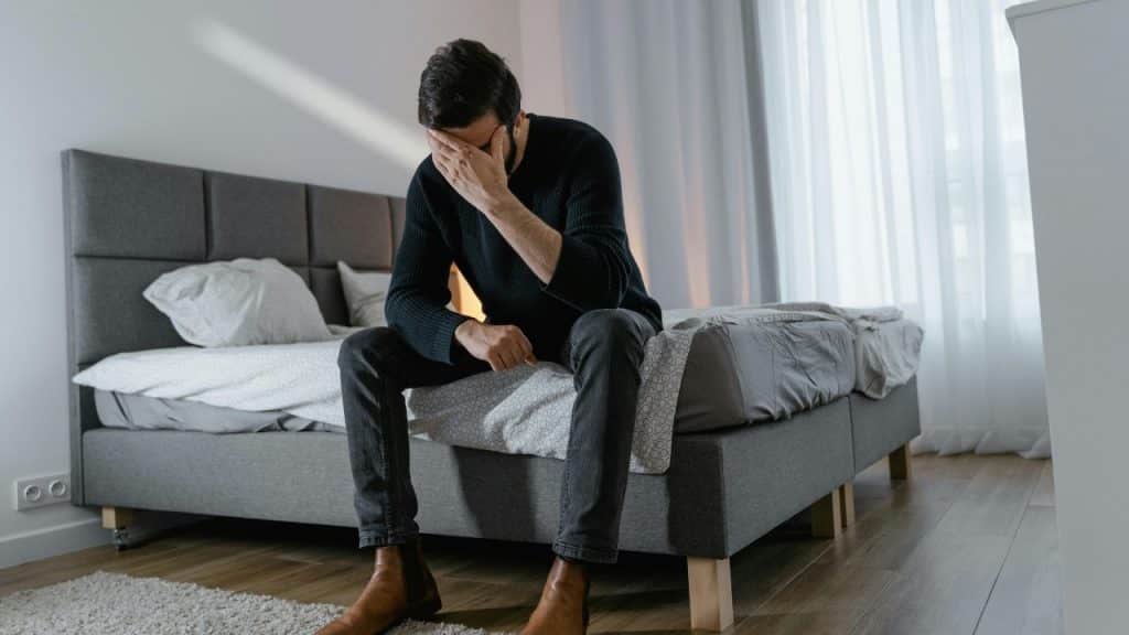 A man sits on the edge of a bed with his face in his hand.