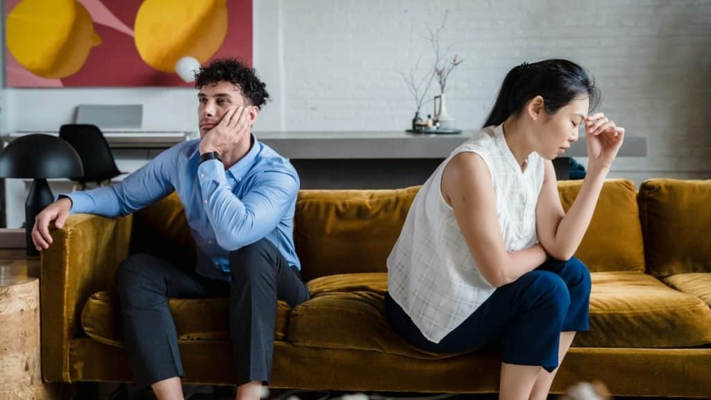 A man and woman sit far apart on a mustard-colored sofa, looking away from each other.