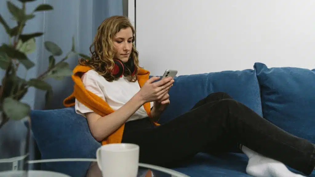 A woman with red headphones around her neck sits on a blue sofa using a smartphone.
