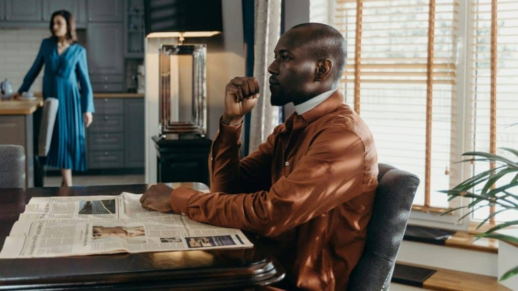 A man reads a newspaper at a table while a woman stands far in the background.