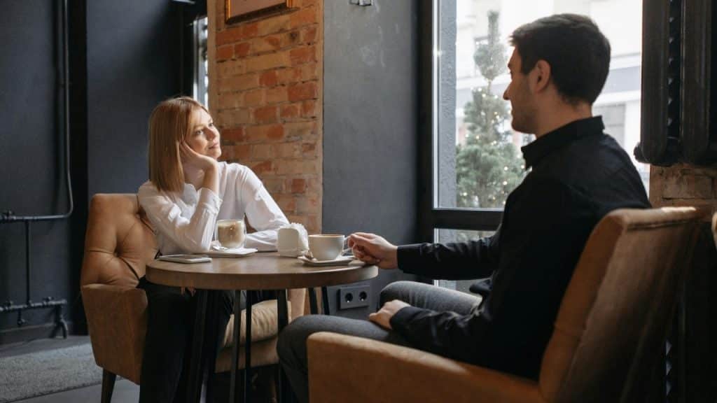 A man and woman sit at a small round table in a cafe with two coffee cups.