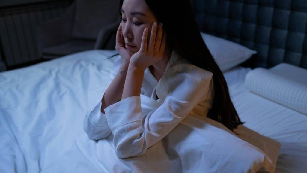 A woman sits on a bed in a dark room with her face resting in her hands.