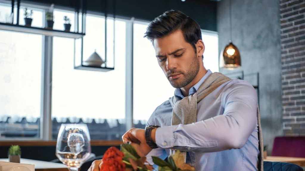 A man in a blue shirt looks down at his wristwatch while sitting at a restaurant.