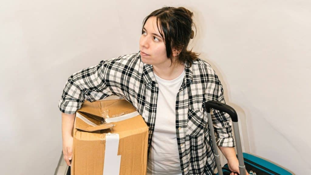 A woman in a flannel shirt holds a cardboard box while standing next to a suitcase.