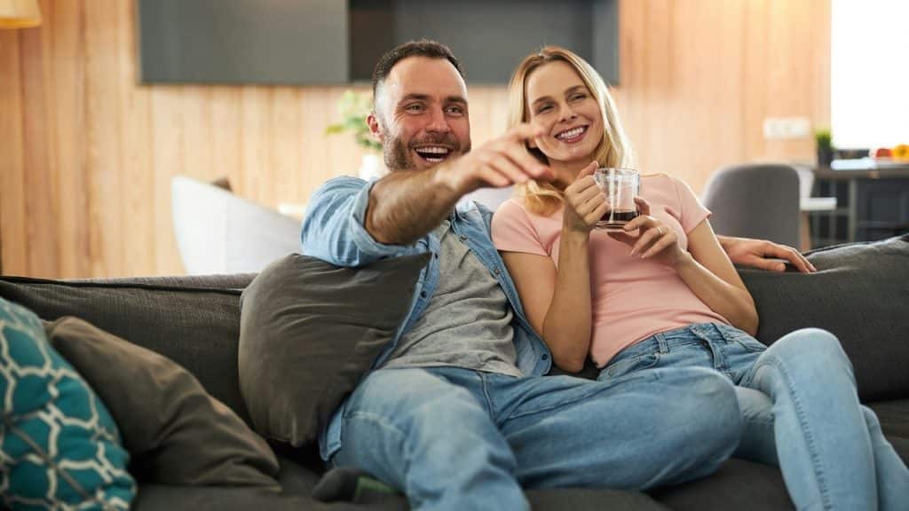 A man and woman sit on a sofa laughing and pointing toward something off-camera in front.