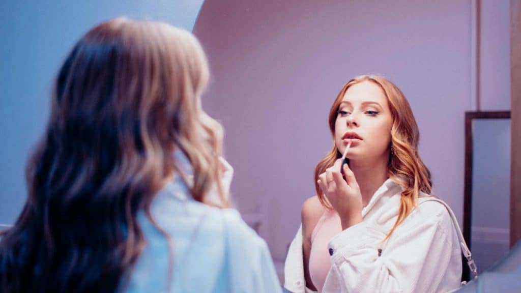 A woman looks into a mirror while applying lip gloss with a small wand and tube.