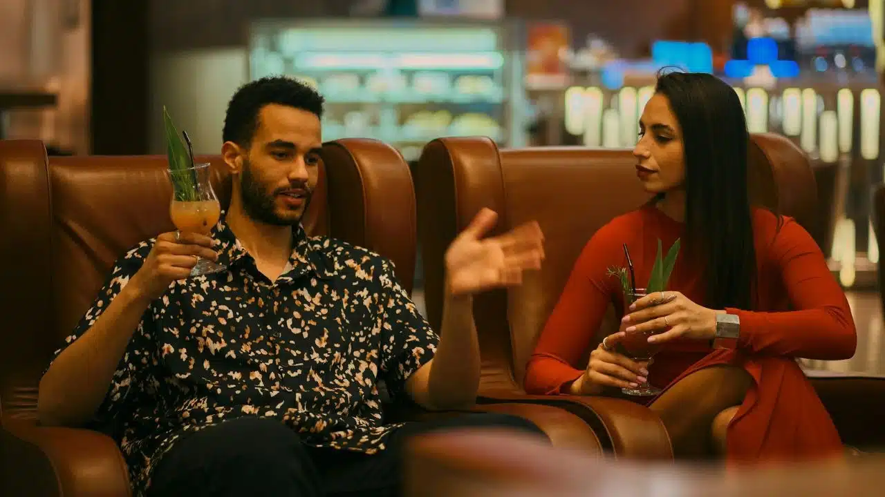A man and woman sit in leather chairs holding cocktails while the man gestures broadly.