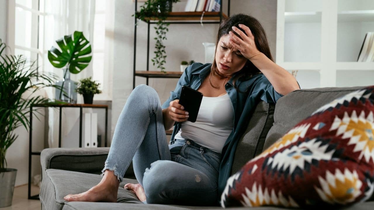 A woman sits on a couch holding a phone with one hand on her forehead.