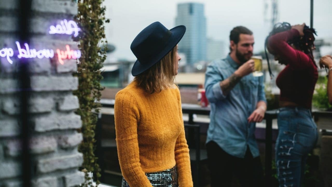 A woman in an orange sweater and blue hat looks away at a rooftop gathering.