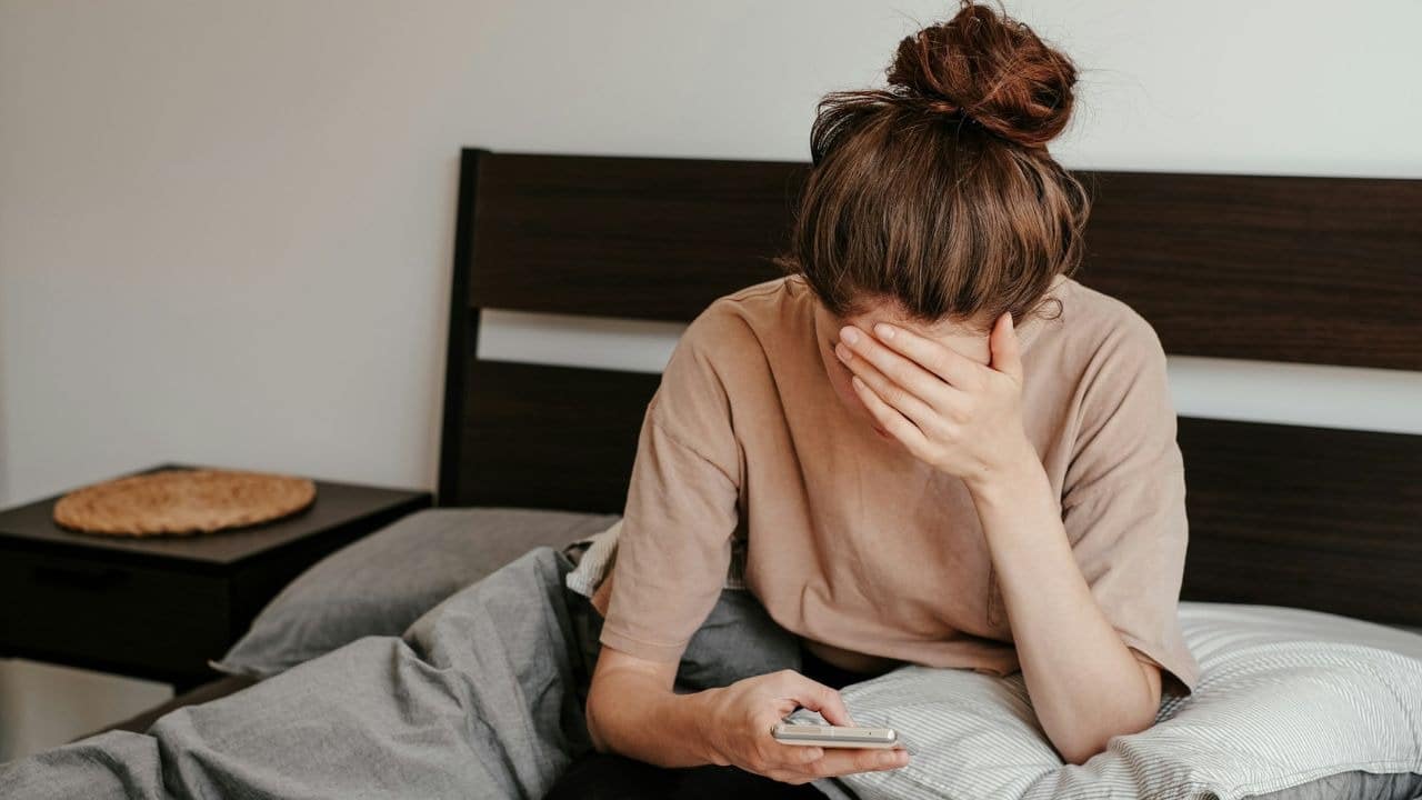 A woman lies on a bed, holding a phone while covering her face with one hand.