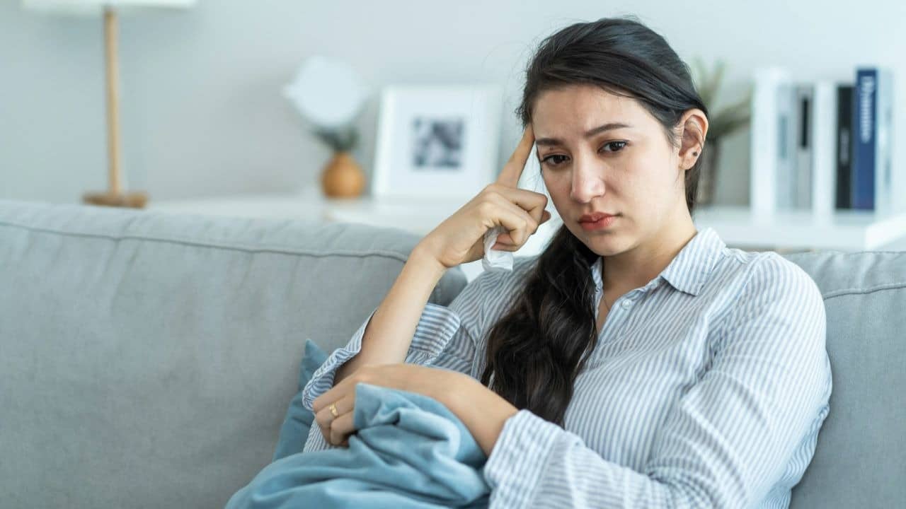 A woman in a striped shirt sits on a couch with a hand to her forehead.
