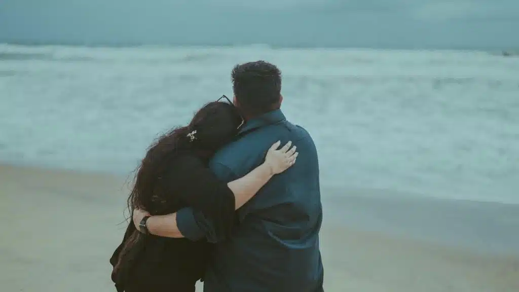 A rearview shot of a couple standing on a sandy beach, looking out at the ocean waves under an overcast sky.