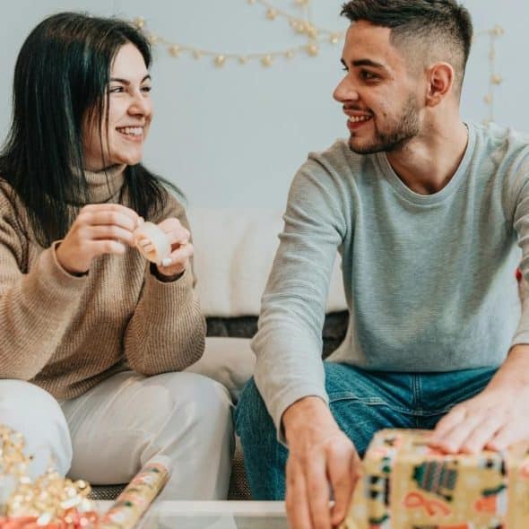 A couple smiling while wrapping gifts together on a couch.