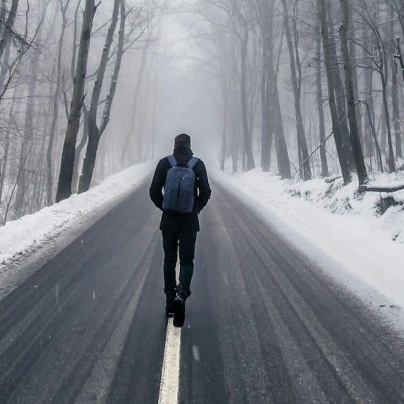 A man wearing a backpack walks on a snowy road with his hands tucked in his pockets.