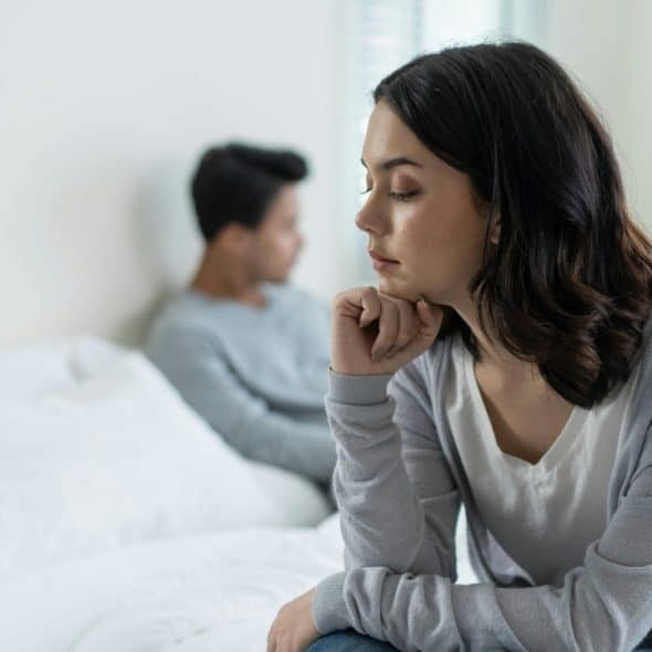 A sad woman sitting on the edge of a bed while her partner lies in it behind her.