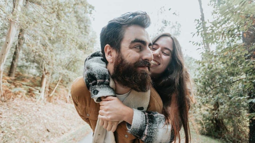 A man with a beard and a brown jacket gives a piggyback ride to a smiling woman with long brown hair along a wooded path.